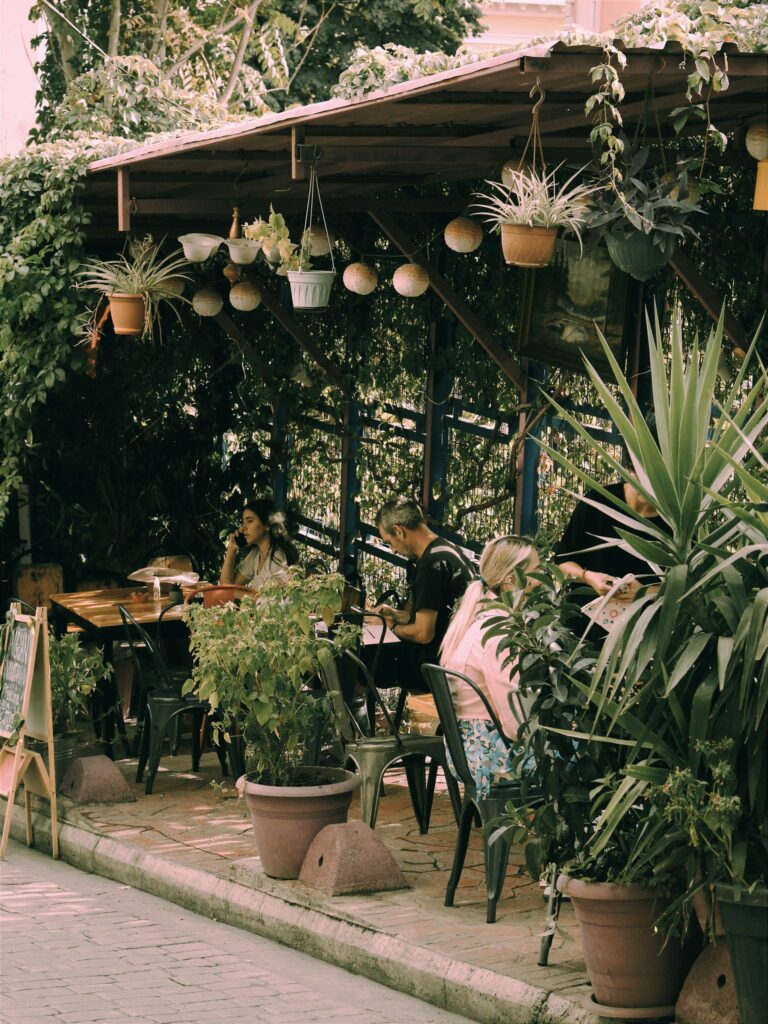 Charming outdoor café with lush plants and patrons enjoying leisurely dining in a garden-like setting.